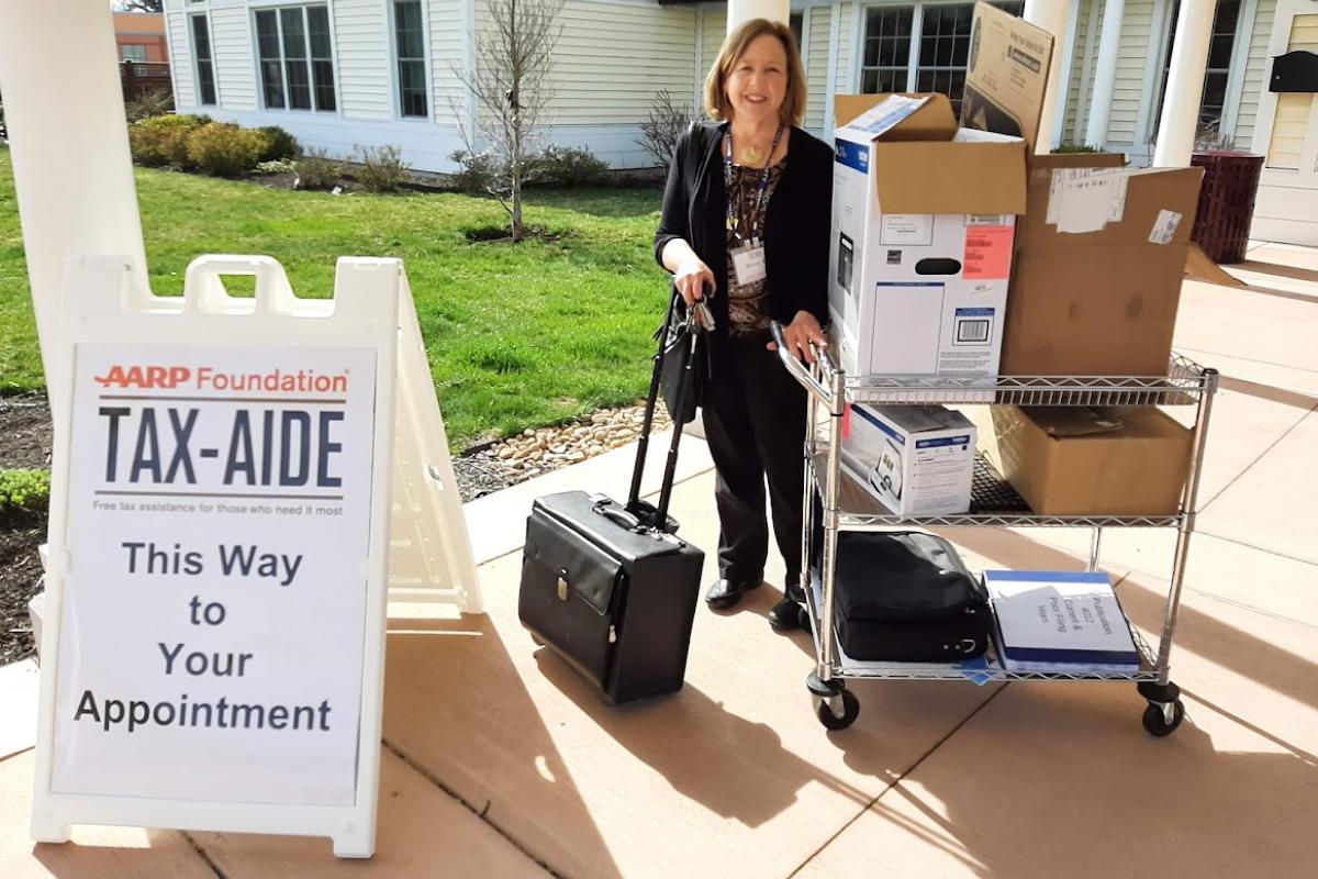 Woman transporting boxes and a suitcase stands next to sandwich board