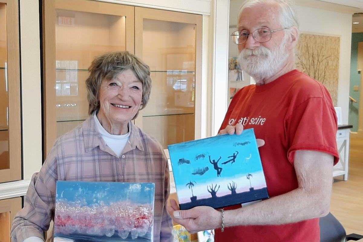 A woman stands next to a man who is holding a small painting