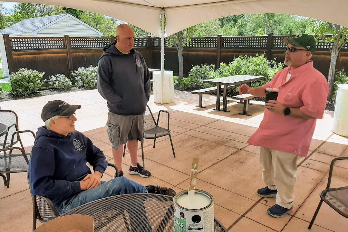 Three men have a conversation under a tent on a patio