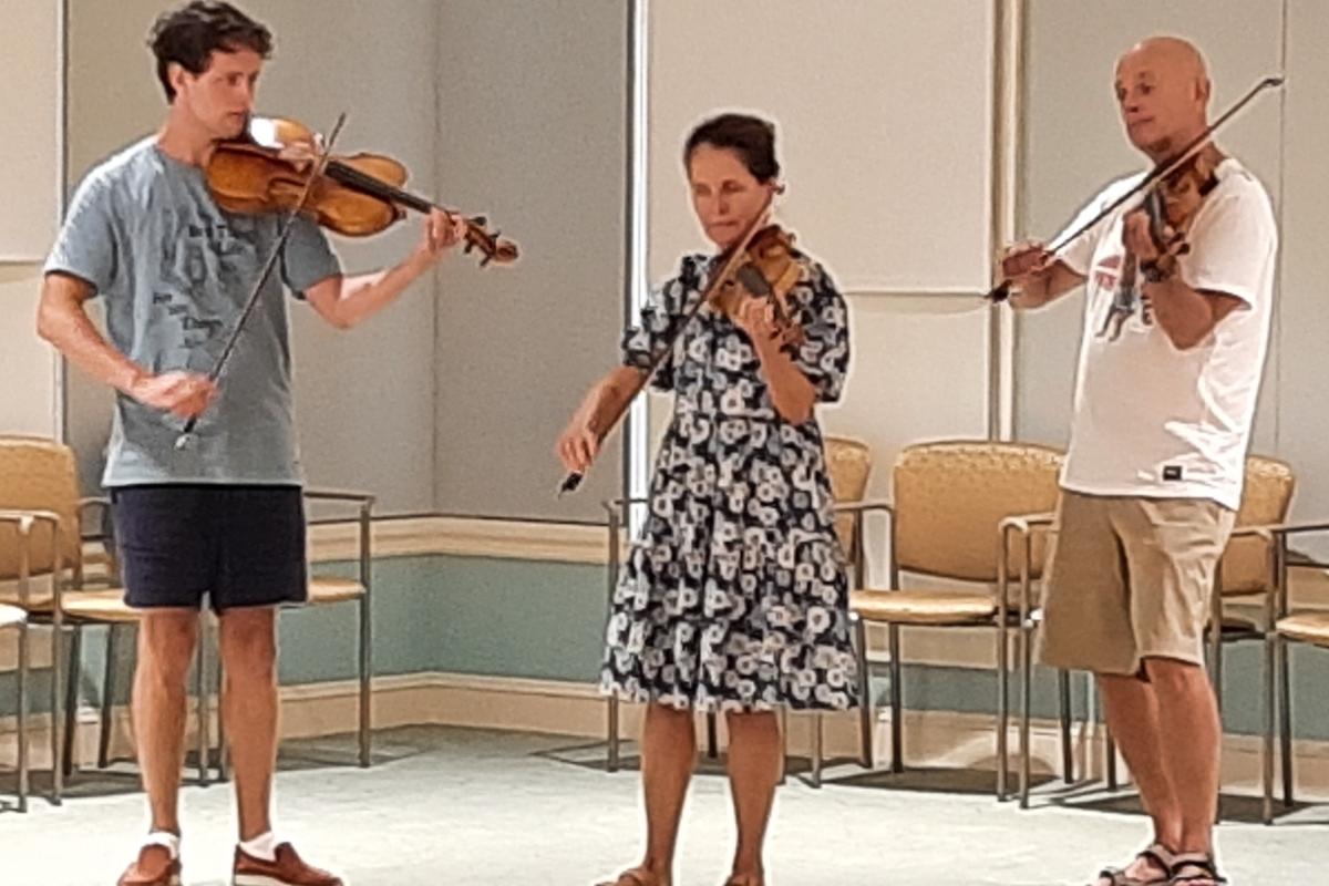 Two men and a women play fiddles on an indoor stage
