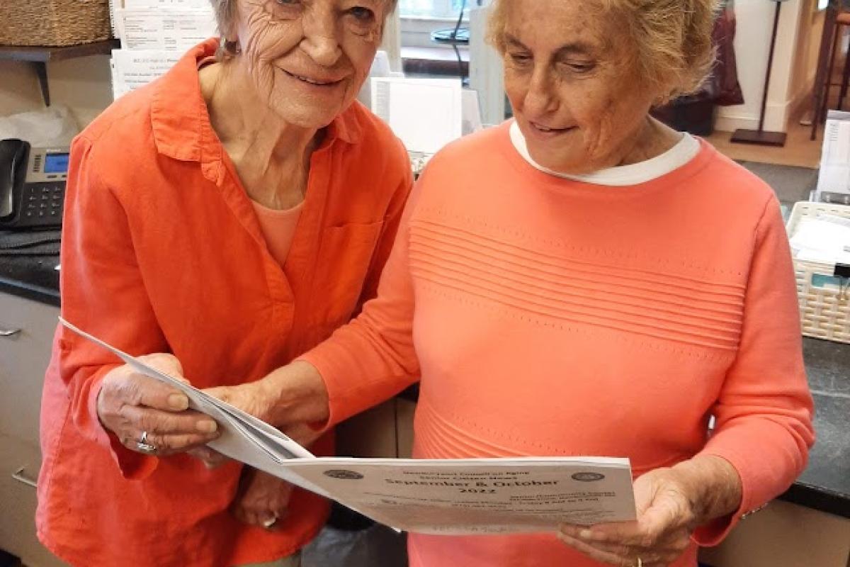 Two women wearingoOrange sweaters look at newsletter together