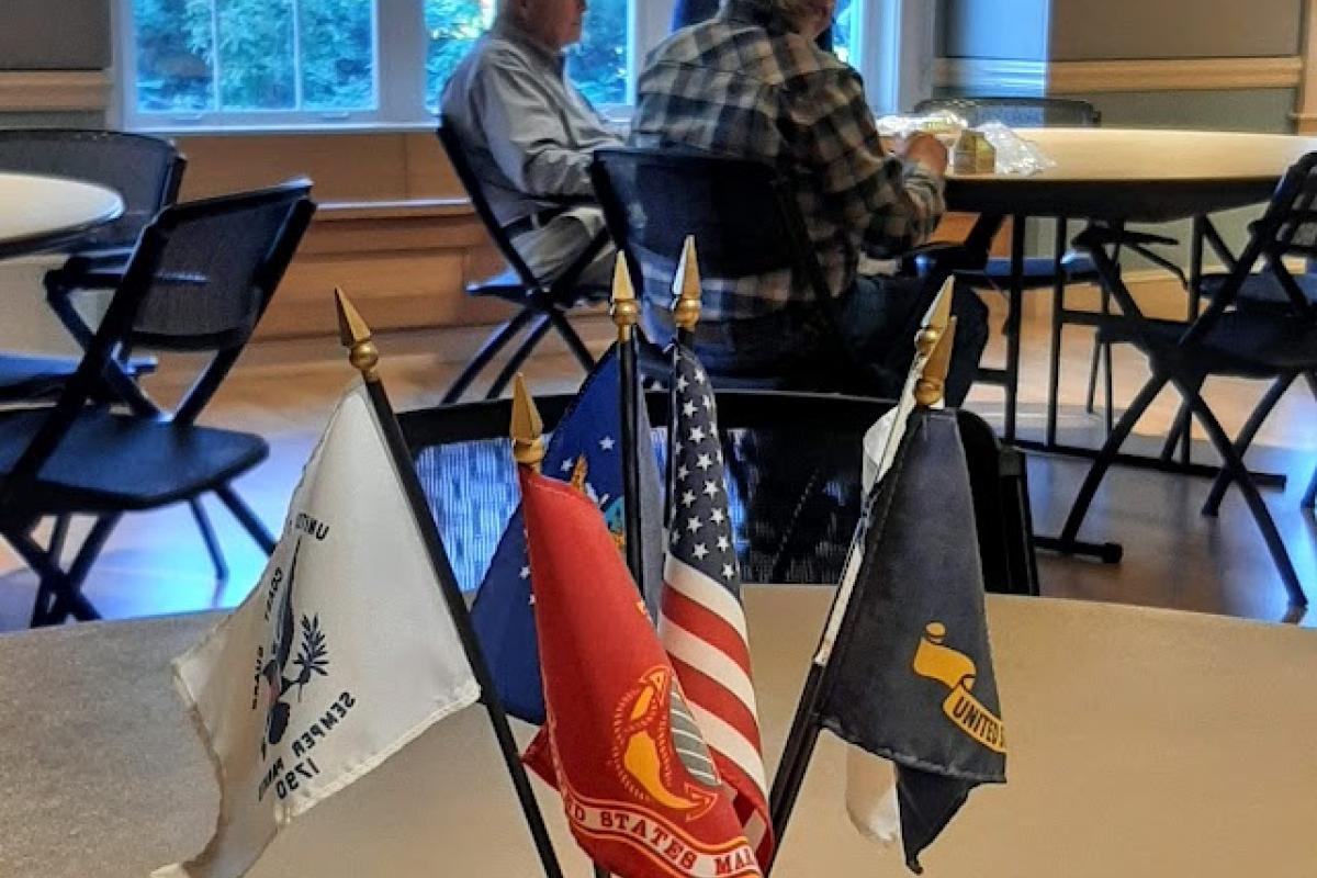 Military flags in front of a man speaking with men sitting 