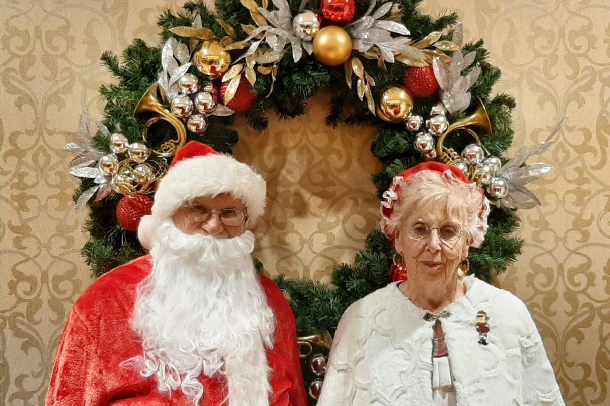 Santa and Mrs Claus Stand in Front of a Large Decorated Wreath