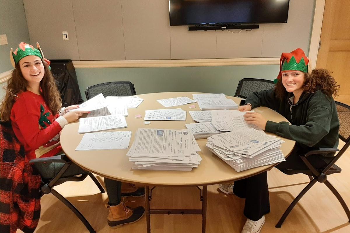 Two young women wearing elf hats assemble a newsletter on a round table