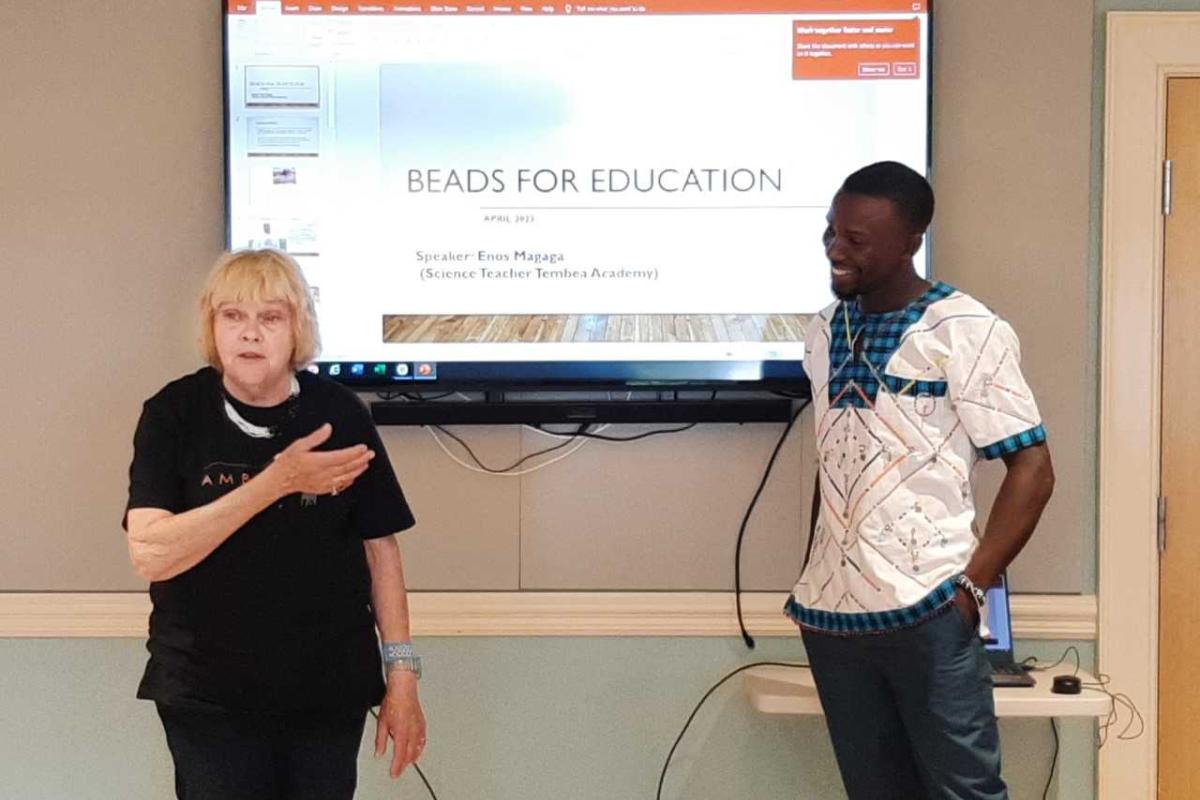 A woman and man stand in front of a projected image that reads "Beads for Education"