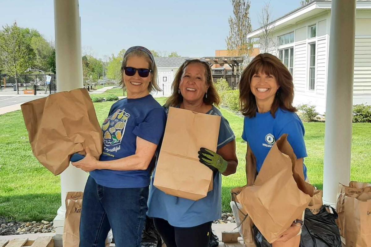 Three smiling women hold packed brown grocery bags as they stand side-by-side outside under a portico
