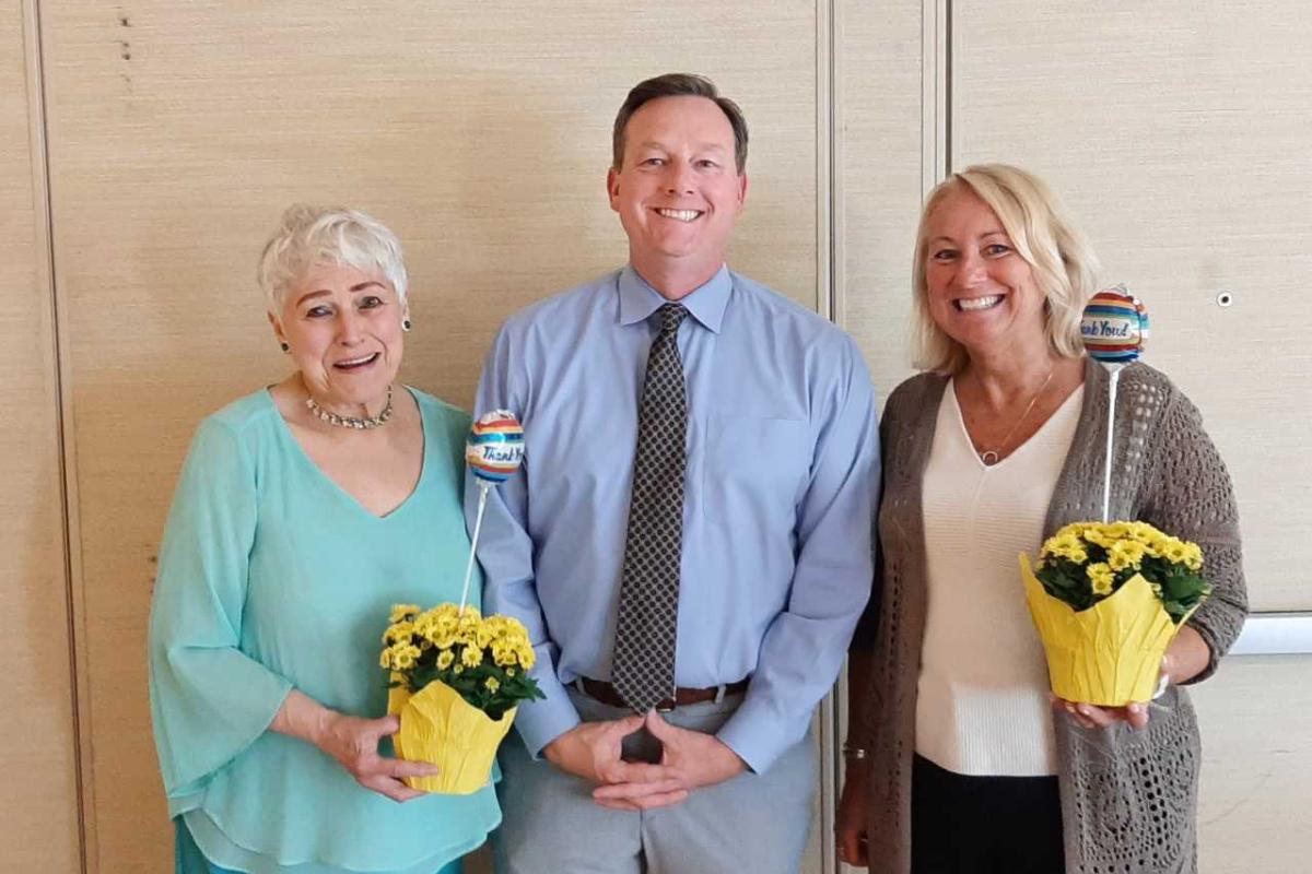 A man poses for a photo between two women who are each holding a plant with bright yellow flowers